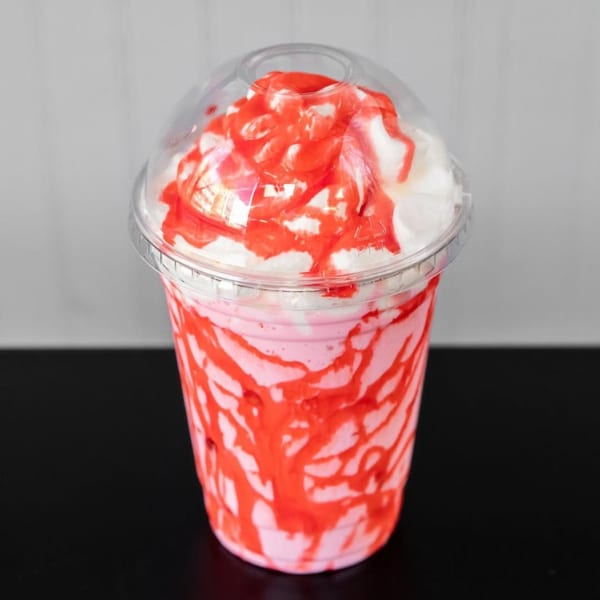 Pink milkshake with whipped cream and strawberry drizzle in a clear plastic cup.