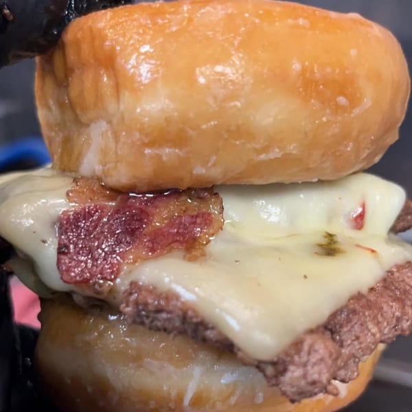 Close-up of a Halal Buns donut burger with melted cheese and bacon, held by a gloved hand.