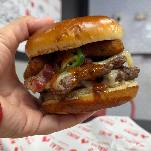 Hand holding a Halal Buns burger with two patties, melted cheese, bacon, jalapeños, and sauce.