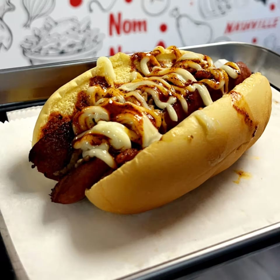 A Halal Buns hot dog with chili, white sauce, and barbecue sauce on a bun.