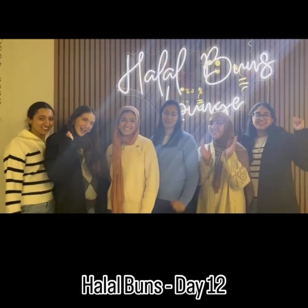 Six young women smiling in front of a neon "Halal Buns" sign.