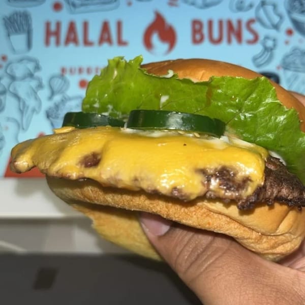 Hand holding a Halal Buns burger with melted cheese, lettuce, and pickles, against a restaurant logo.