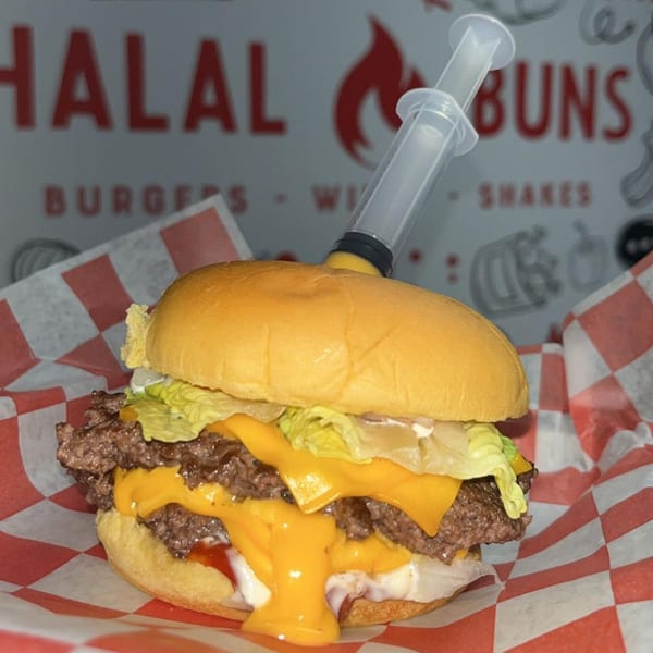 A Halal Buns double cheeseburger with lettuce, sauce, and a syringe injecting cheese, on checkered paper.